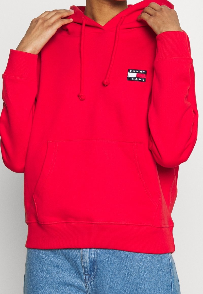 Personne ajustant la capuche d'un sweat à capuche Tommy Jeans rouge vif porté avec un jean en denim bleu.