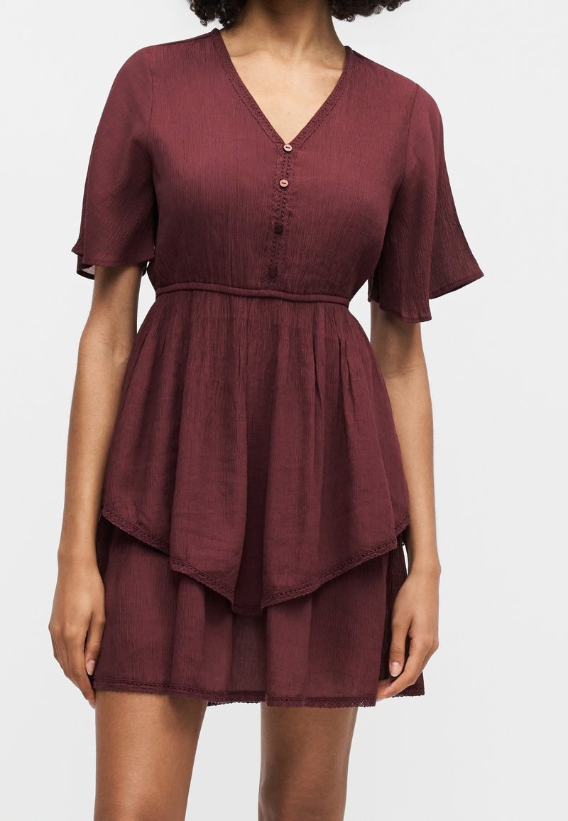 Femme portant une robe courte bordeaux à étages avec un col en V, des manches courtes volantées et trois boutons sur le corsage avant.