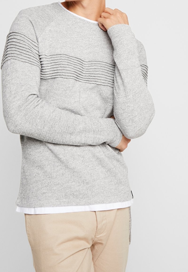 Personne portant un pull gris clair avec une texture côtelée horizontale sur la poitrine et le haut des manches, un pantalon beige et un t-shirt blanc en dessous.
