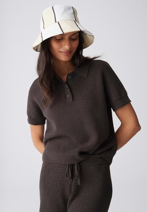 Frau trägt ein dunkles, gestricktes Poloshirt mit kurzen Ärmeln und passende Kordelzughosen, kombiniert mit einem weißen Bucket Hat mit schwarzen Streifen.