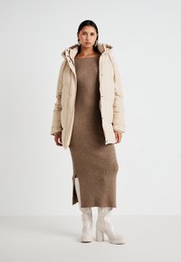 Doudoune beige avec capuche, associée à une robe marron côtelée avec une fente sur le côté. Le modèle porte des bottes blanches jusqu'au genou.