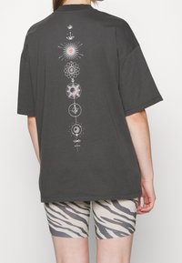 Persona vestida con una camiseta de gran tamaño gris oscuro con un diseño celestial vertical y pantalones cortos de ciclista con estampado de cebra beige, vista desde la espalda.