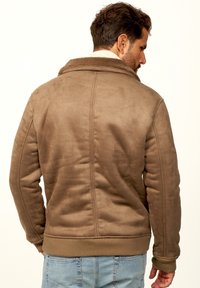 Braune Kunstwildlederjacke mit einem plüschigen Kragen, gerippten Bündchen und Saum sowie dekorativen Nähten; getragen über hellblauen Jeans.