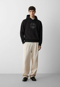 Felpa nera con cappuccio e logo, abbinata a pantaloni crema larghi e scarpe nere. Tessuto liscio e silhouette rilassata.
