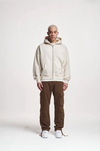 Hoodie a zip in un beige chiaro con grandi tasche frontali, indossato su pantaloni cargo marroni con più tasche, abbinato a sneakers bianche.