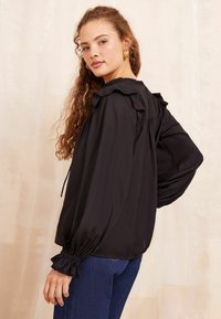 Blusa nera con accentuazioni arricciate sulle spalle e sui polsini, caratterizzata da un fit comodo e maniche lunghe, realizzata in un tessuto morbido e leggero.