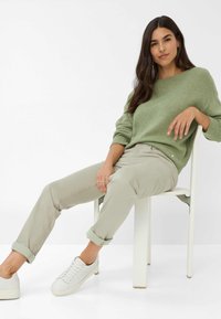 Pull en tricot vert, pantalon beige clair et baskets blanches. Le pull a une coupe décontractée, des manches retroussées et une texture visible.