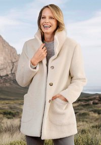 Cremefarbener Fleecepullover mit hohem Kragen, drei Knöpfen und Seitentaschen. Weiche Textur, lockere Passform, getragen über einem grauen Pullover.