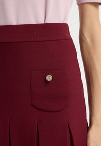Burgundy kjol med en texturerad tygkvalitet, utrustad med en liten fackficka med en silverknapp och en midjeband. Designad med vertikala veck.