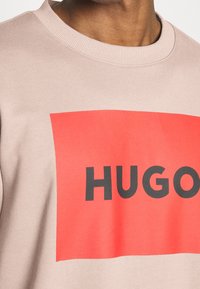 Sudadera Hugo en tejido beige claro que presenta un gran gráfico rectangular rojo con la palabra "HUGO" en letras negras en negrita.