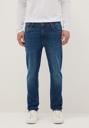 Hombre vestido con vaqueros ajustados azules, camisa blanca de manga larga y zapatillas blancas, de pie frente a un fondo liso.