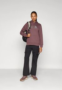 Sweatshirt com capuz roxa com logótipo, calças cargo pretas e sapatilhas castanhas. Modelo segura uma mochila preta. Design simples, roupa casual para atividades ao ar livre.