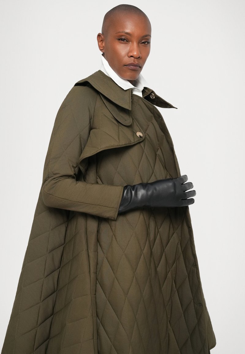 Femme avec la tête rasée portant un manteau olive matelassé, une chemise blanche et des gants en cuir noirs, regardant la caméra avec confiance.