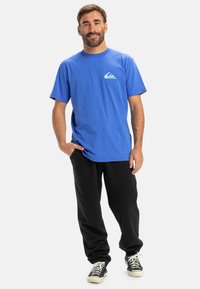 Modrá bavlnená tričko s malým svetlomodrým logom na ľavom prsníku, skombinované s čiernymi teplákmi a čiernymi teniskami. Jednoduchý, pohodlný strih.