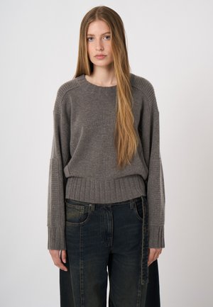 Junge Frau mit langen, glatten Haaren, die einen grauen Strickpullover und dunkle, locker sitzende Jeans trägt, vor einem schlichten hellen Hintergrund stehend.