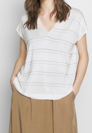 Mujer con suéter blanco de manga corta con cuello en V y finas rayas horizontales, y pantalones beige de pierna ancha, con las manos en los bolsillos.