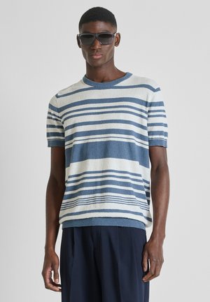 Hombre con grandes gafas de sol negras, camisa de manga corta a rayas azules y blancas, y pantalones azul marino oscuro, de pie frente a un fondo liso.