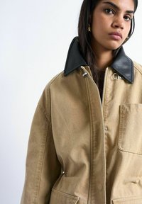 Jeune femme aux longs cheveux bruns portant un manteau beige avec un col en cuir noir, se tenant devant un fond clair uni.