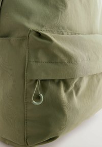 Mochila de tela verde con una superficie texturizada, gran bolsillo frontal con un cierre curvado, y sutiles detalles de costura.