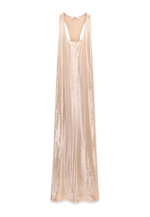Femme aux longs cheveux blonds dans une robe beige sequins dos nu, portant des bracelets dorés et des talons beiges, debout devant un fond clair uni.