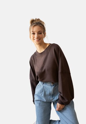 Młoda kobieta uśmiechająca się, ubrana w ciemnobrązową, oversize'ową bluzę i jasnoniebieskie dżinsy z wysokim stanem, pozująca na jednolitym jasnym tle.