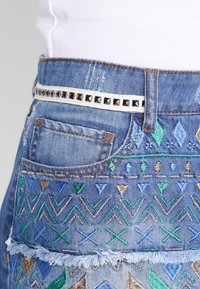 Jupe en denim ornée de broderies multicolores en vert, bleu et or, ourlet effiloché et ceinture blanche avec des clous noirs.