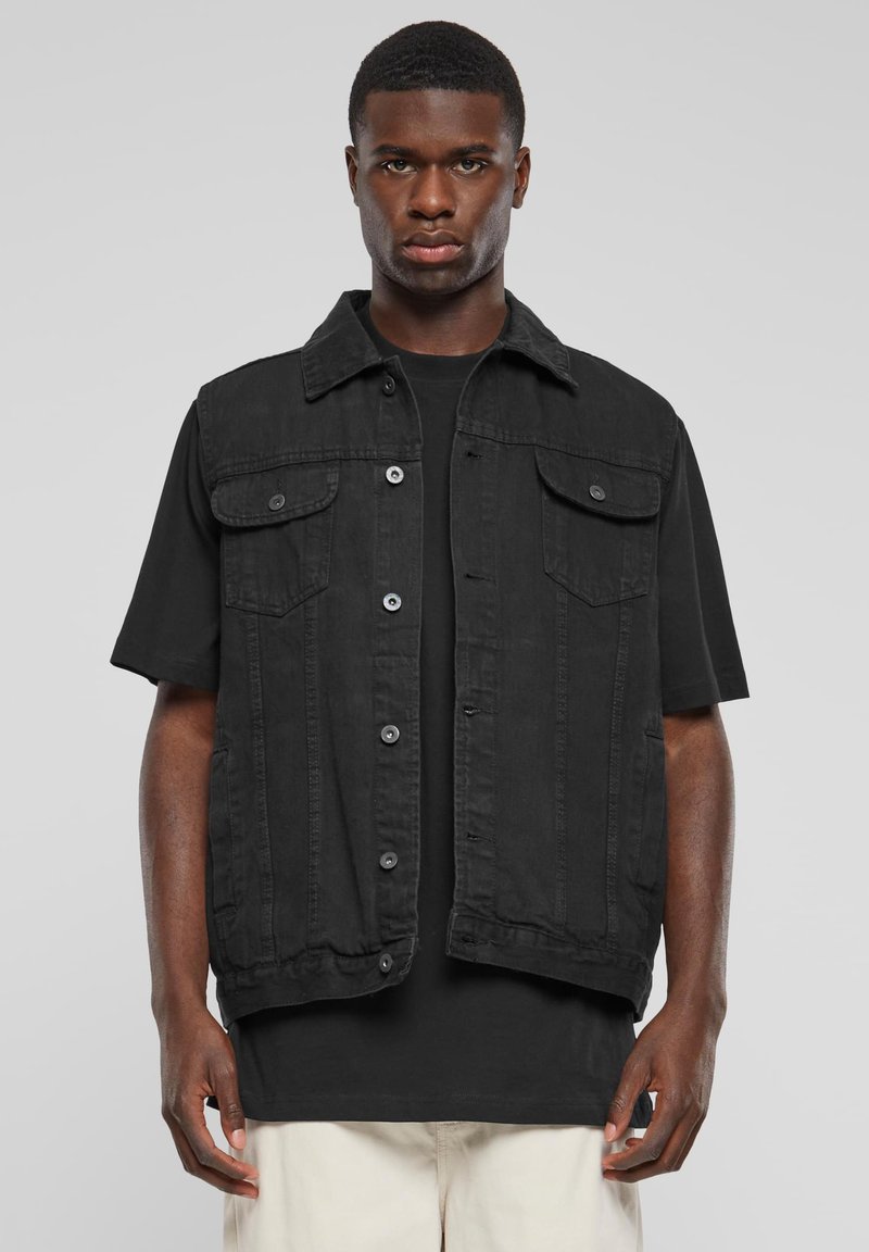 Jeune homme aux cheveux courts portant un gilet en denim noir boutonné sur un t-shirt noir et un pantalon de couleur claire, debout devant un fond uni.