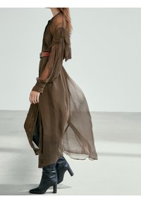 Lång, olivgrön genomskinlig klänning med plisserad textur, prydda ärmar och ett brunt bälte. Matchad med svarta högklackade stövlar.