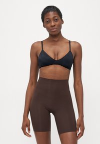Vrouw met kort haar, gekleed in een zwarte bralette en hoge bruine compressiekorte broek, staande voor een effen witte achtergrond.