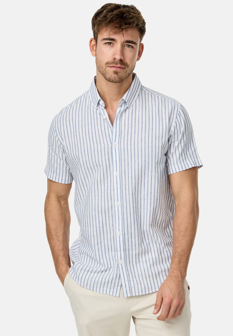 Homme portant une chemise blanche à manches courtes avec des rayures verticales bleues et un pantalon beige, debout avec une main dans la poche devant un fond uni.