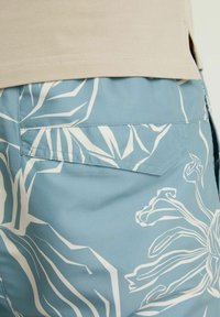 Gros plan sur un short bleu à motif floral abstrait blanc et un ourlet de chemise beige qui chevauche la taille.
