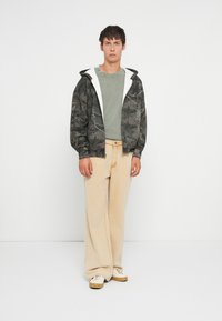 Hooded jacket v tmavom maskáčovom vzore s šedými a bielymi akcentmi, skombinovaný so zeleným tričkom a béžovými širokými nohavicami.