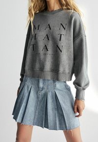Grijs cropped sweatshirt met zwarte tekst "MANHATTAN" en "NEW YORK CITY" eronder, gecombineerd met een lichtblauwe geplooide denimrok met een rafelrand.