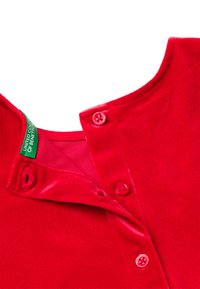 Czerwona aksamitna koszula z okrągłym dekoltem, dwoma widocznymi czerwonymi guzikami oraz zieloną metką z logo wewnątrz kołnierzyka. Gładka tekstura.