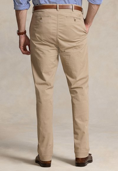 Mężczyzna w beżowych spodniach, brązowym skórzanym pasku, niebieskiej koszuli z długim rękawem podwiniętym, brązowym skórzanym zegarku i brązowych eleganckich butach, stojący z rozluźnionymi rękami.