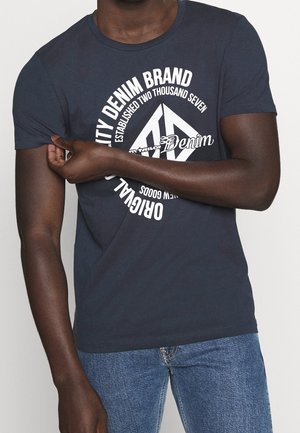 Personne portant un t-shirt bleu foncé avec un texte circulaire blanc "Original City Denim Brand" et un jean bleu, ajustant la manche du bras gauche.