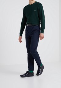Ciemnozielony sweter z dzianiny, ciemnoniebieskie spodnie typu tapered, czarne sneakersy i zielone skarpetki; posiada logo na klatce piersiowej oraz pasek.