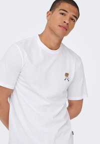 White cotton t-shirt with short sleeves featuring a small black and yellow embroidered design of a character carrying stacked rings.
