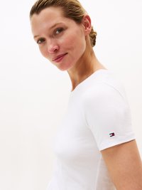 T-shirt bianco in cotone con maniche corte, design aderente, presenta un piccolo logo rosso, bianco e blu sulla manica sinistra.