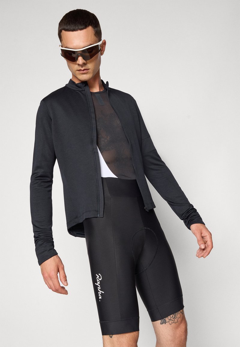 Männer Radbekleidung bestehend aus einer leichten schwarzen Jacke, einem Mesh-Oberteil und schwarzen gepolsterten Shorts mit dem weißen "Rapha."-Logo an der Seite.
