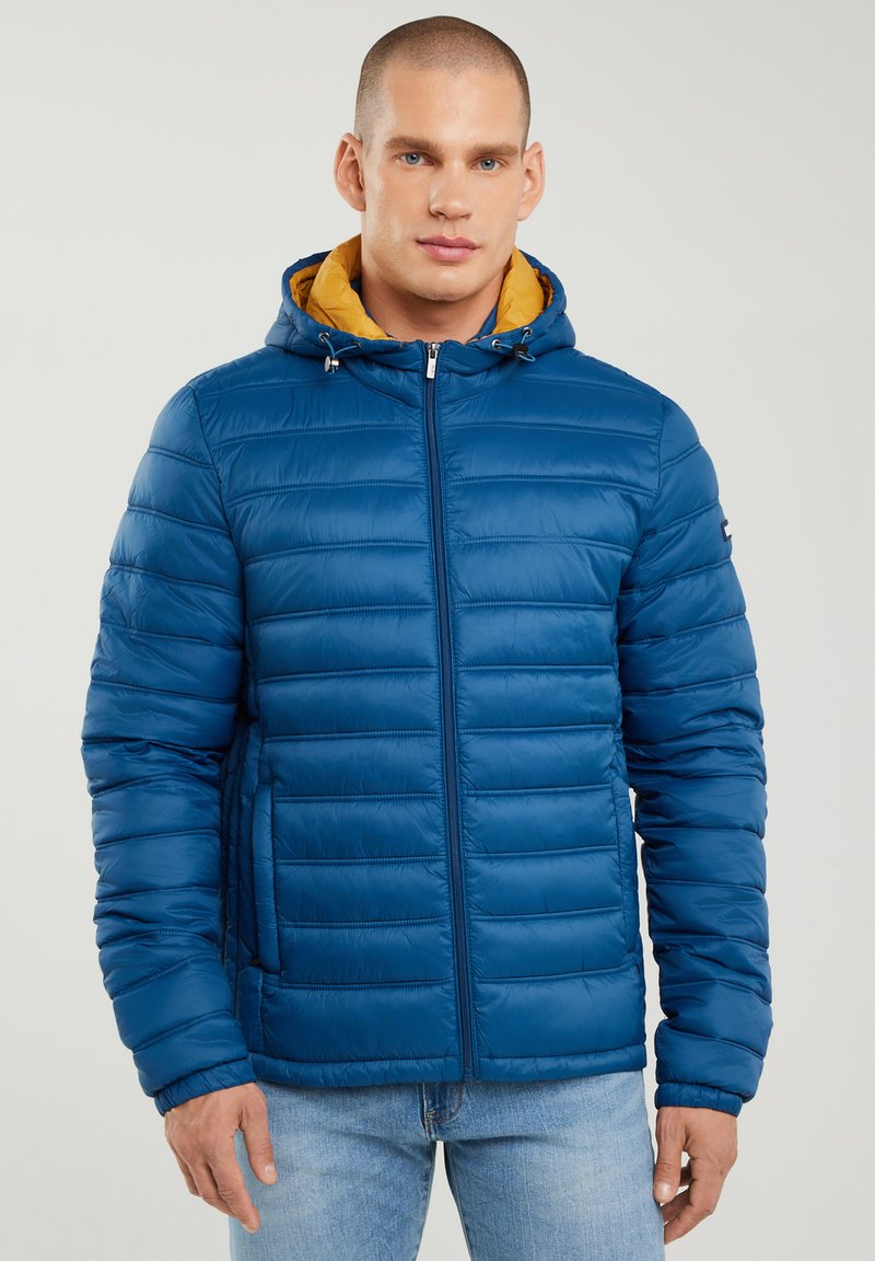 Mann trägt eine blaue gesteppte Steppjacke mit gelbem Kapuzenfutter, steht vor einem einfarbigen hellen Hintergrund.