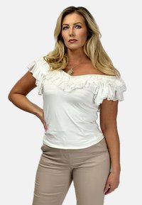 Biała bluzka hiszpanka z falbanami, wykonana z gładkiego materiału. Połączona z dopasowanymi beżowymi spodniami, podkreślającymi smukłą sylwetkę.