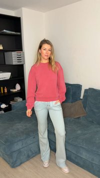 Sweat-shirt rouge avec poignets côtelés, associé à un pantalon large à carreaux bleu clair. En arrière-plan, une étagère sombre avec divers objets.