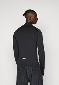 Bluză neagră cu mâneci lungi, fabricată dintr-un material texturat, cu guler înalt și un branding subtil la tiv. Asortarea cu pantaloni negri completează ținuta.