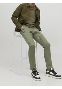 Olivgrön jacka och t-shirt, kombinerat med ljusgröna byxor. Avslutat med svarta och vita sneakers. Man sitter på en vit barstol.