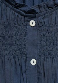 Mörkblå skjorta med smockdetaljer, rund halsringning och två vita knappar. Har mjuk tygtextur och samlad sömnad.