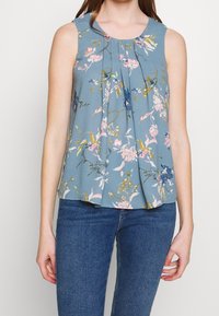 Kukkaistyyppinen hihaton pusero vaaleansinisessä kankaassa, jossa on taitoksilla varustettuja yksityiskohtia. Sisältää värikkäitä kukkakuoseja ja pyöreän pääntien, yhdistettynä farkkuihin.