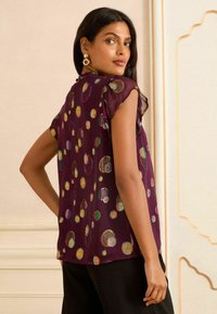 Blusa in plum trasparente con maniche corte, caratterizzata da motivi circolari colorati e un colletto rotondo. Texture liscia con dettagli vivaci.
