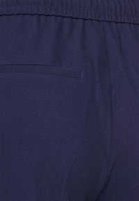 Námorně modré kalhoty vyrobené z hladké tkaniny, se jednoduchým elastickým pasem a malou zadní kapsou se skrytým otvorem.