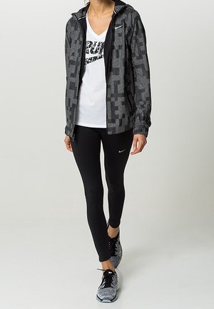 Completo sportivo Nike in bianco e nero, composto da una giacca con zip a motivo, una maglietta bianca con logo, leggings neri e sneakers grigie.